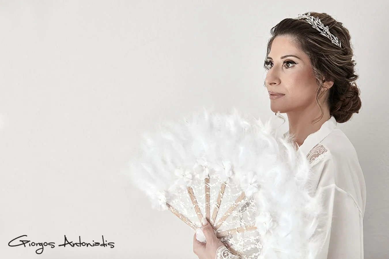 A woman with dark hair styled in an updo, wearing a tiara, and holding a white feathered fan, standing against a plain light background.