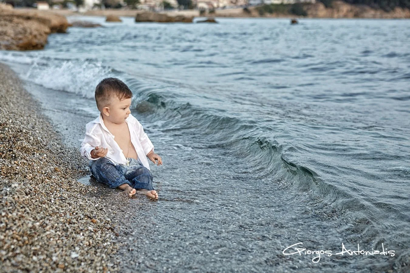 Young boy sitting on a pebbled beach, wearing a white shirt and jeans, eyes focused on the water as small waves lap at his feet.