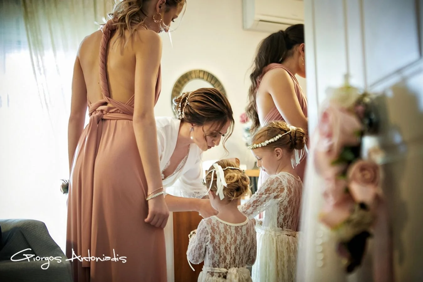 Women and girls dressed for a wedding, with one woman helping a young girl with her dress in a brightly lit room with flowers and decorations.
