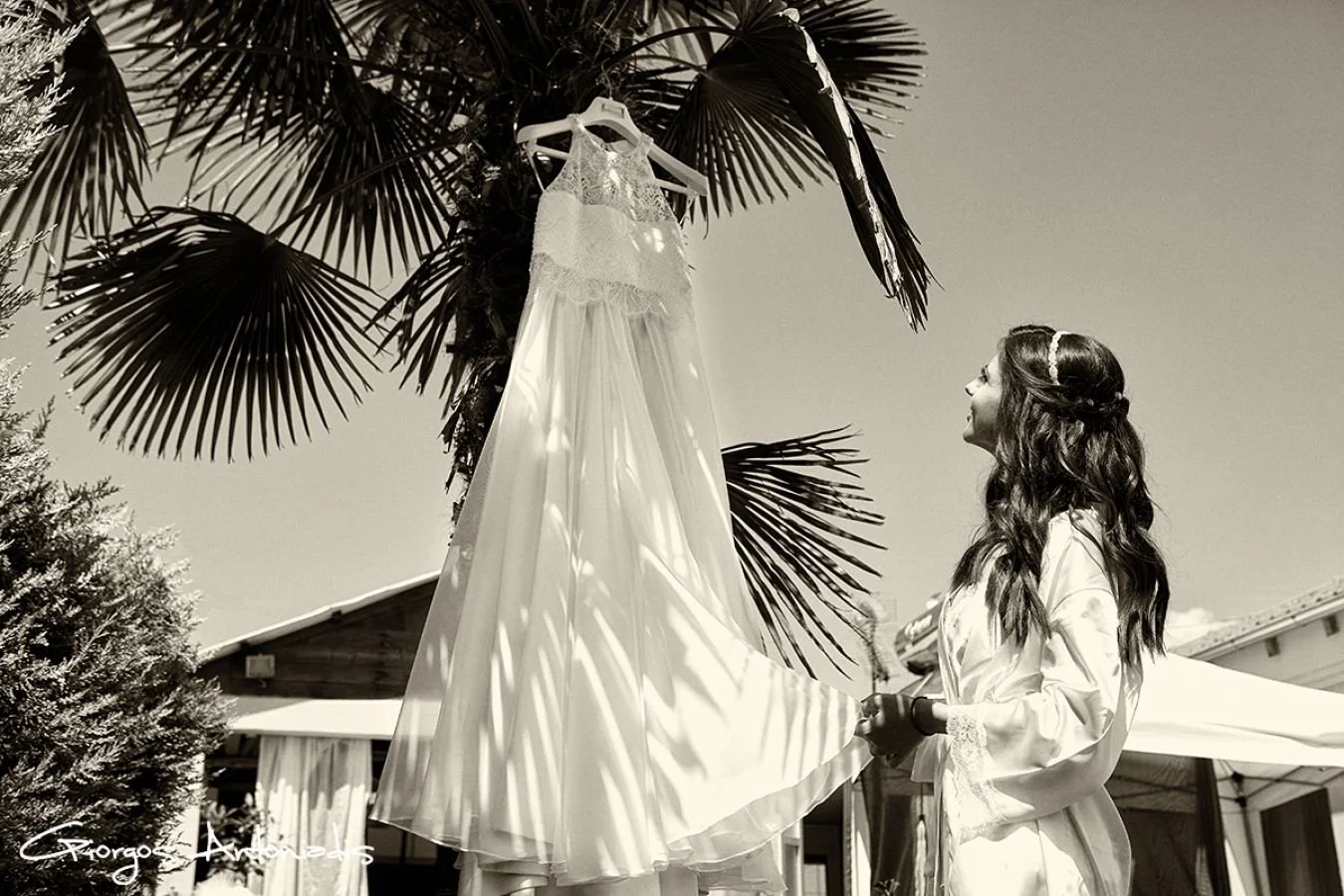A woman with long wavy hair, wearing a satin robe, looks up happily at a wedding dress hanging from a palm tree, outdoors on a sunny day.