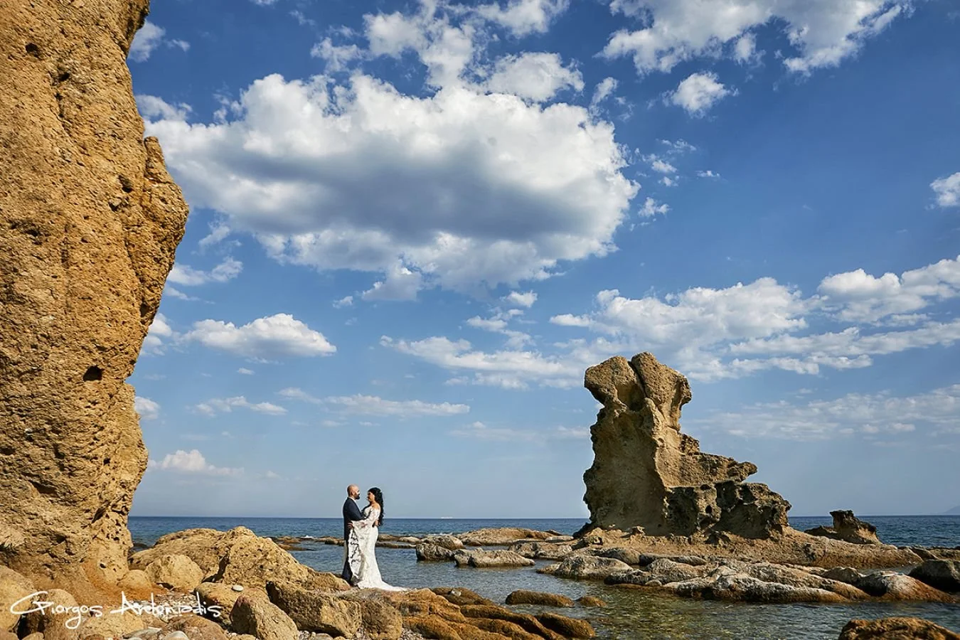 A couple dressed in wedding attire standing on rocky shore by the ocean, with large rock formations and a partly cloudy sky in the background.