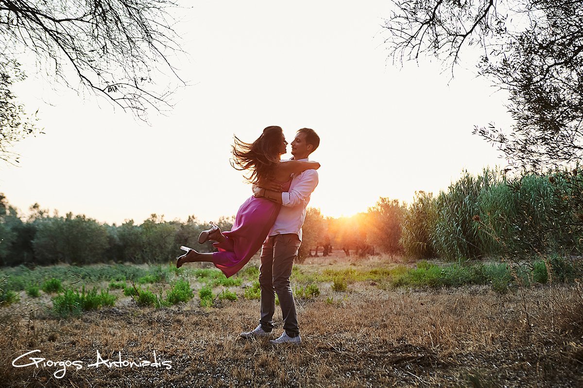 A man lifting a woman in a purple dress during sunset in a natural outdoor setting with trees and grassy fields.