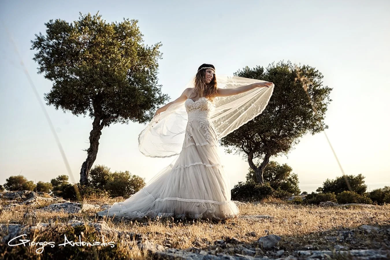 A woman in a flowing white wedding dress standing on rocky ground outdoors with two trees in the background, holding her arms out to the sides as her veil flows behind her in the wind.