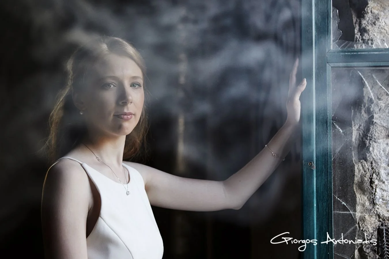 A young woman with red hair in a white dress standing by a glass window with smudges, with a rocky background visible through the glass.