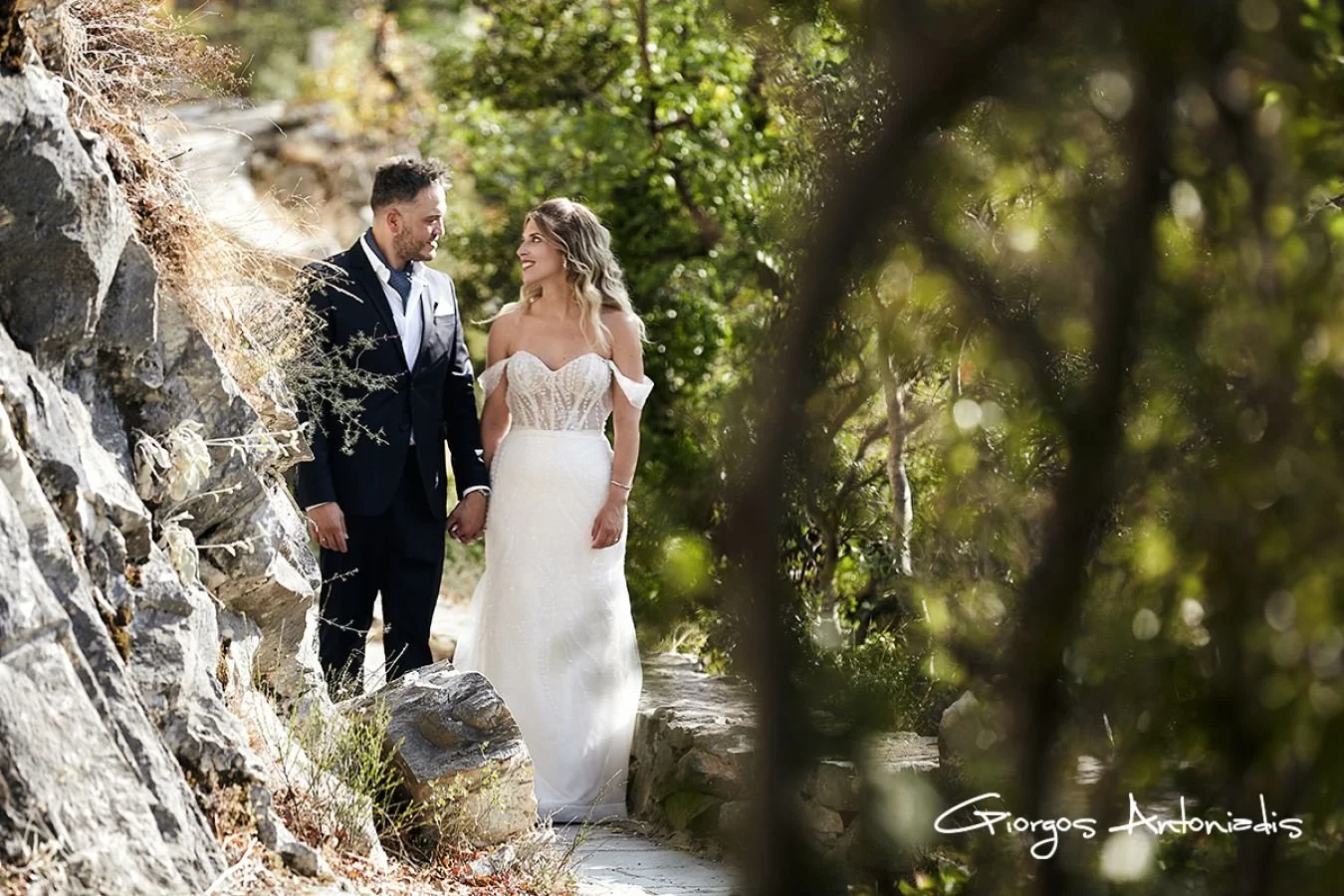 A bride and groom holding hands and smiling at each other outdoors surrounded by rocks and trees.