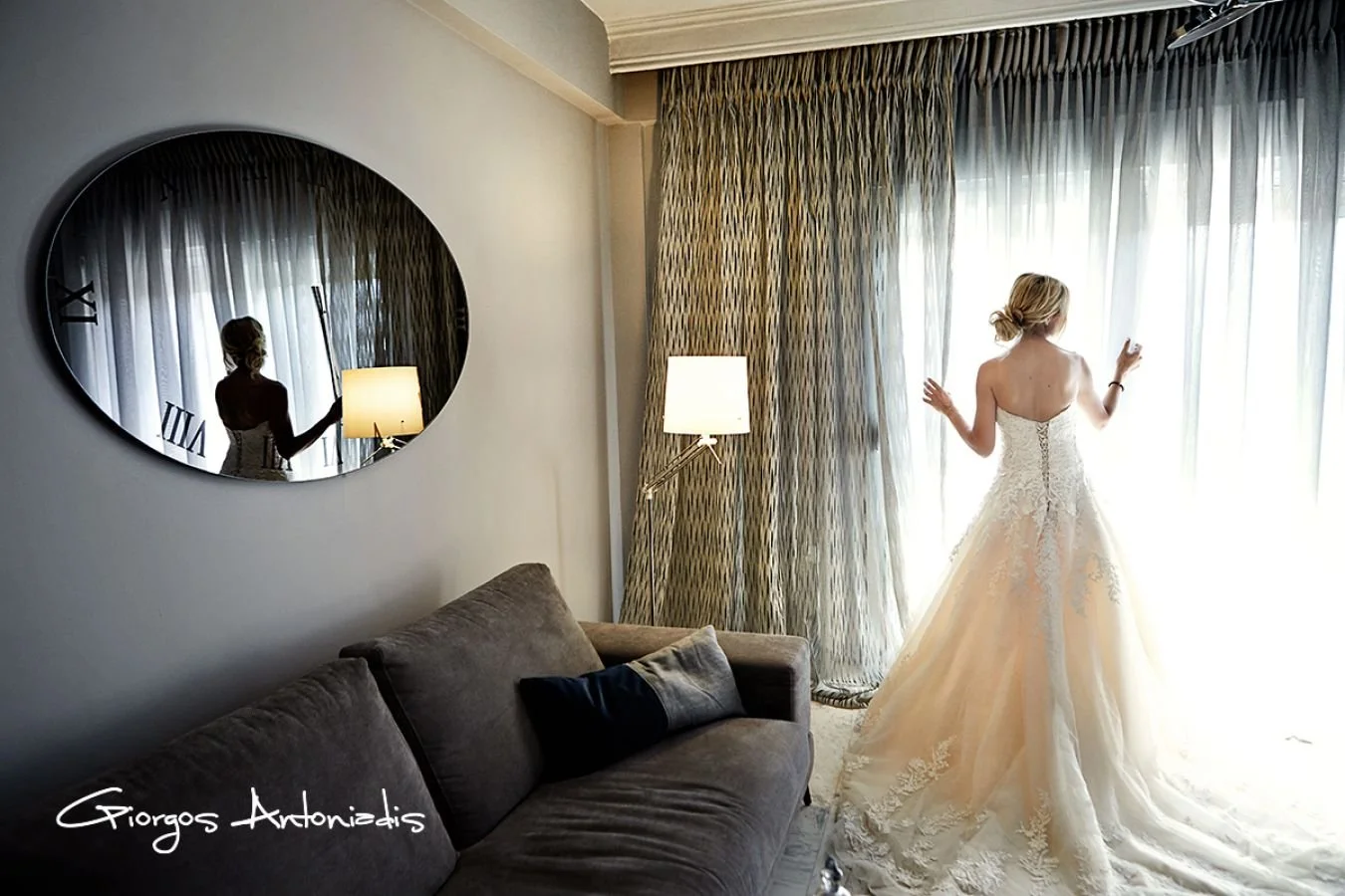A woman in a wedding dress looking out a large window with curtains, seen from behind. Her reflection is visible in a round mirror on the wall.
