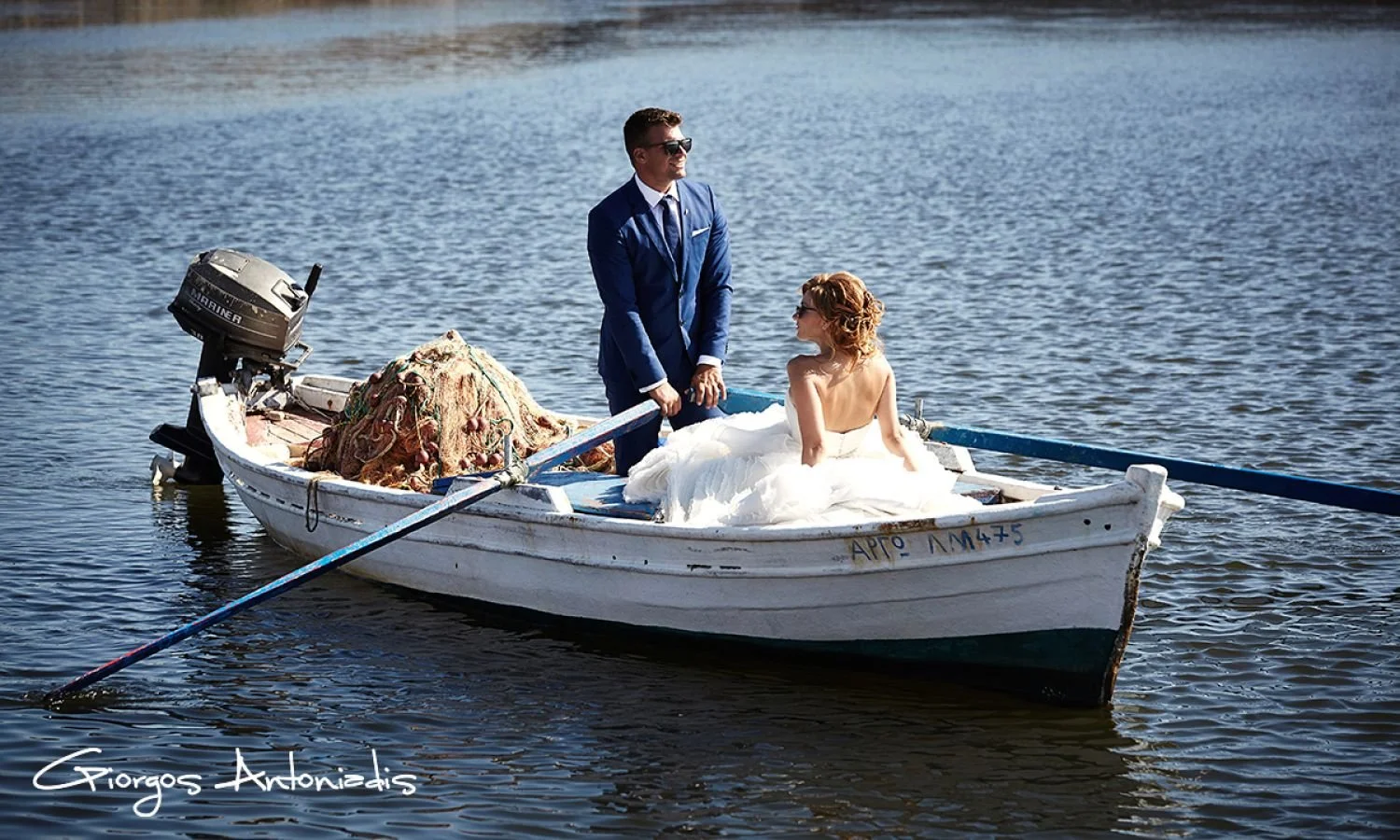 A man in a blue suit and woman in a white wedding dress on a small boat on water, with the woman sitting and the man standing, surrounded by fishing equipment and a small outboard motor.