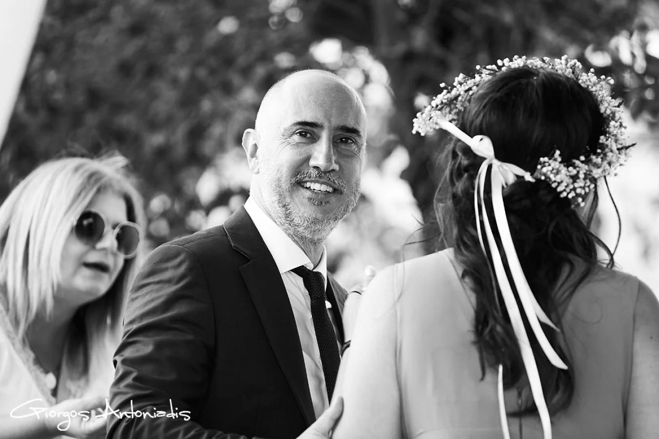 Black and white photo of a man in a suit smiling at a woman with a flower crown, and a woman with sunglasses in the background.