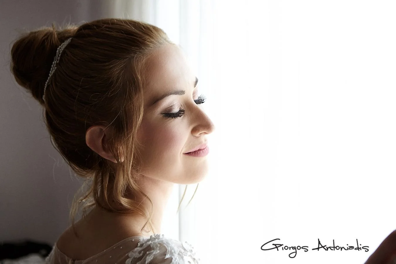 A young woman with red hair styled in an updo, wearing a white lace garment, standing near a window with her eyes closed and a gentle smile.