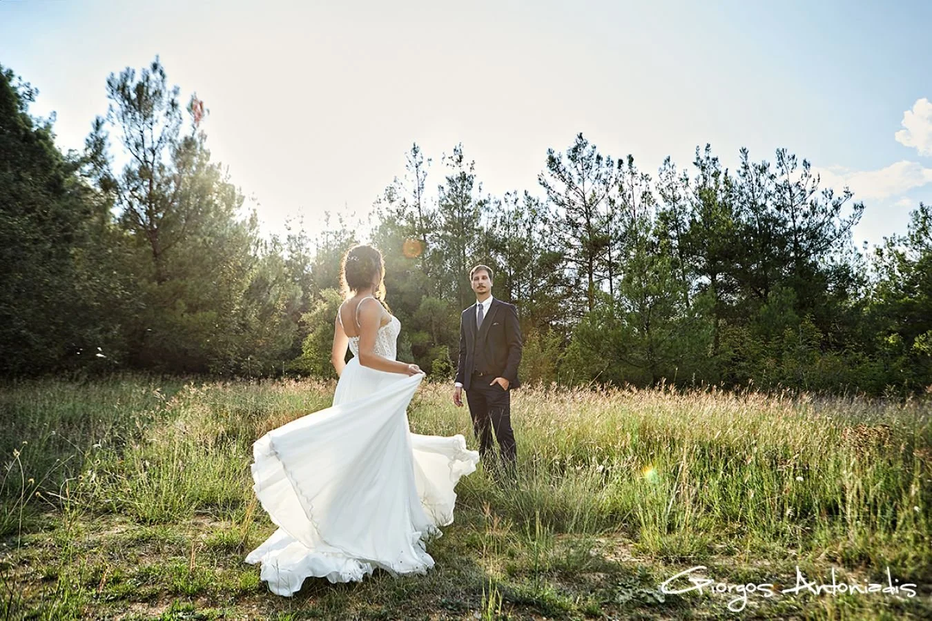 A bride and groom standing in a grassy field with trees in the background during sunset.