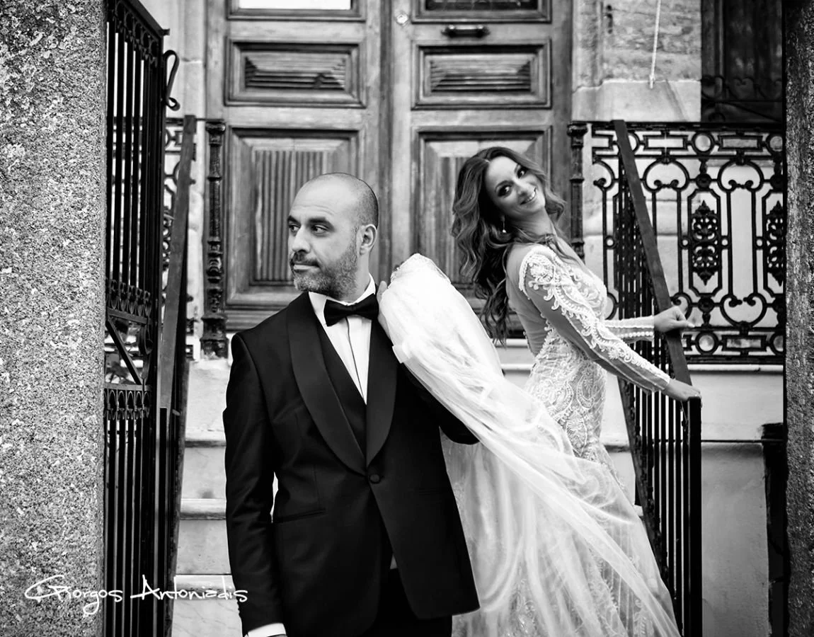 A black and white photo of a groom in a tuxedo and a bride in a lace wedding dress posing outside a building with wooden doors and decorative iron railings.