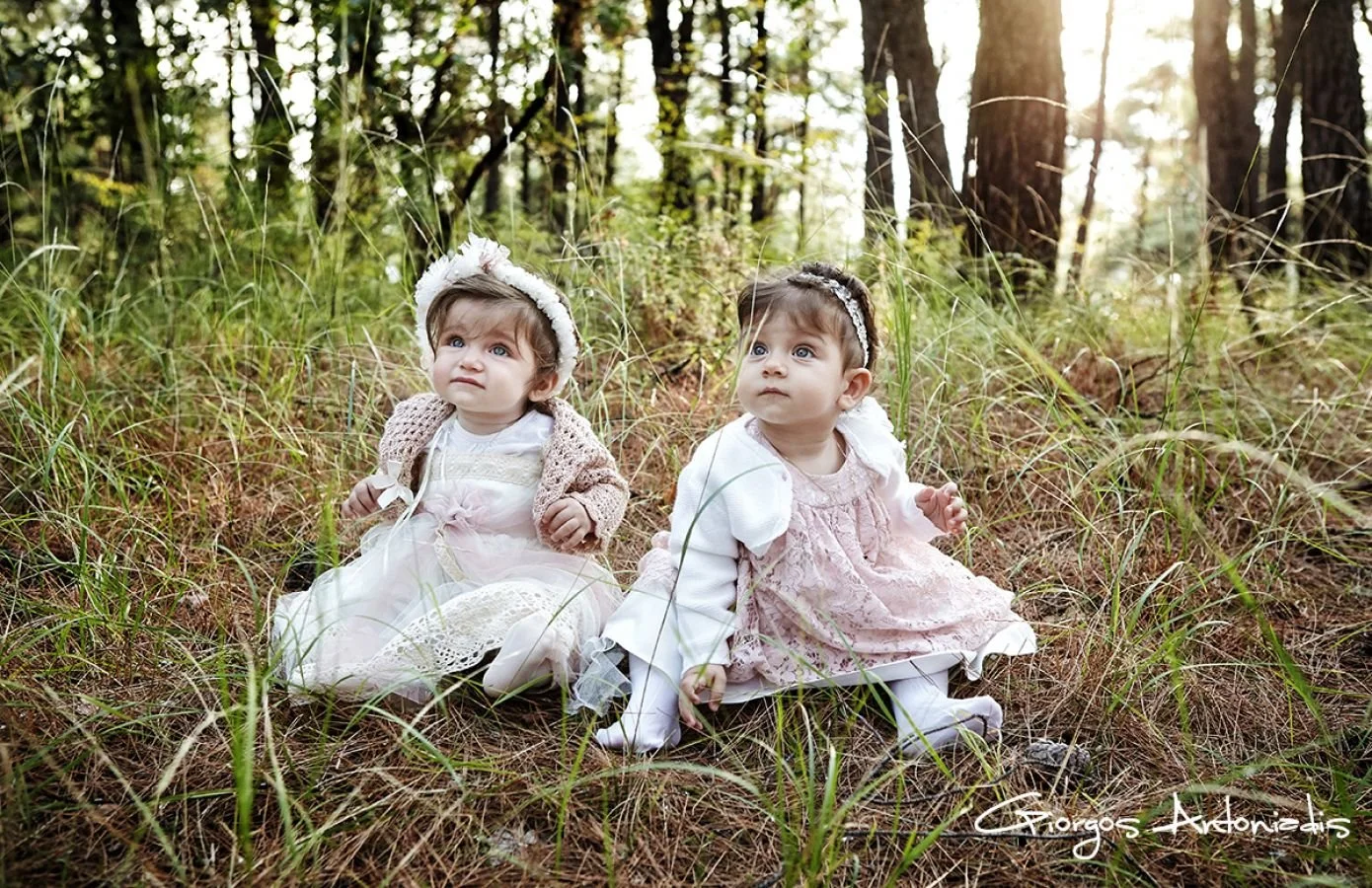 Two young girls sitting in a forest clearing, wearing white dresses and headbands, surrounded by tall grass and trees, with sunlight filtering through the trees behind them.