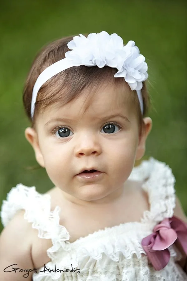 Close-up of a young girl with blue eyes wearing a white headband with large white flowers and a white dress with ruffled straps and a pink bow, outdoors with a green background.