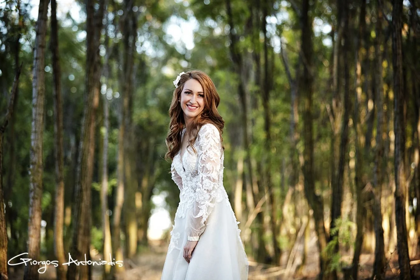 A smiling woman in a white lace wedding dress standing in a forest with tall trees and green foliage, sunlight filtering through the trees.