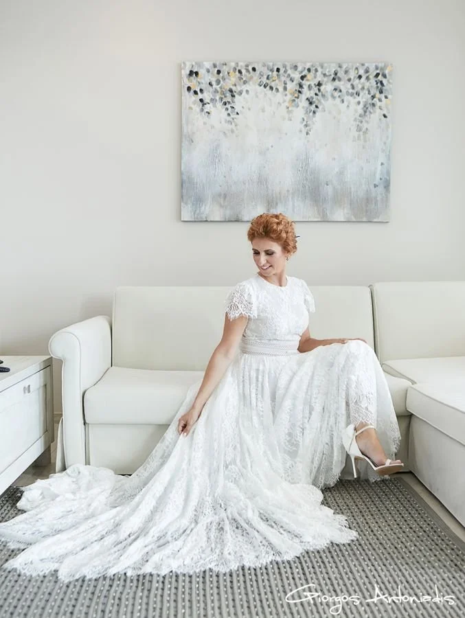 A woman with red curly hair in a white lace wedding dress sitting on a white couch in a modern room, adjusting her dress and wearing high heeled shoes.