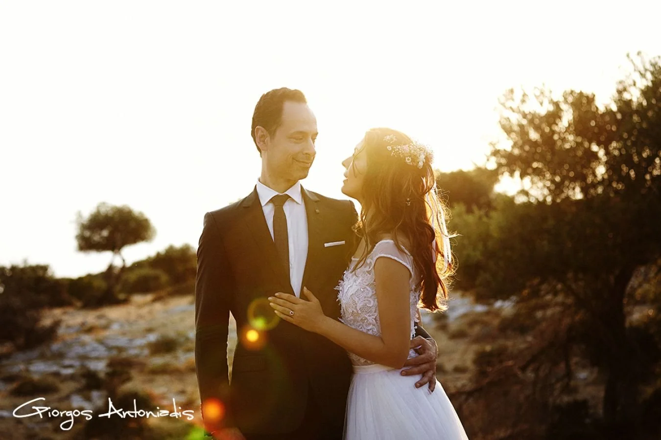 A bride and groom embrace outdoors at sunset, with the bride wearing a white wedding dress and the groom in a black suit, in a natural setting with trees.
