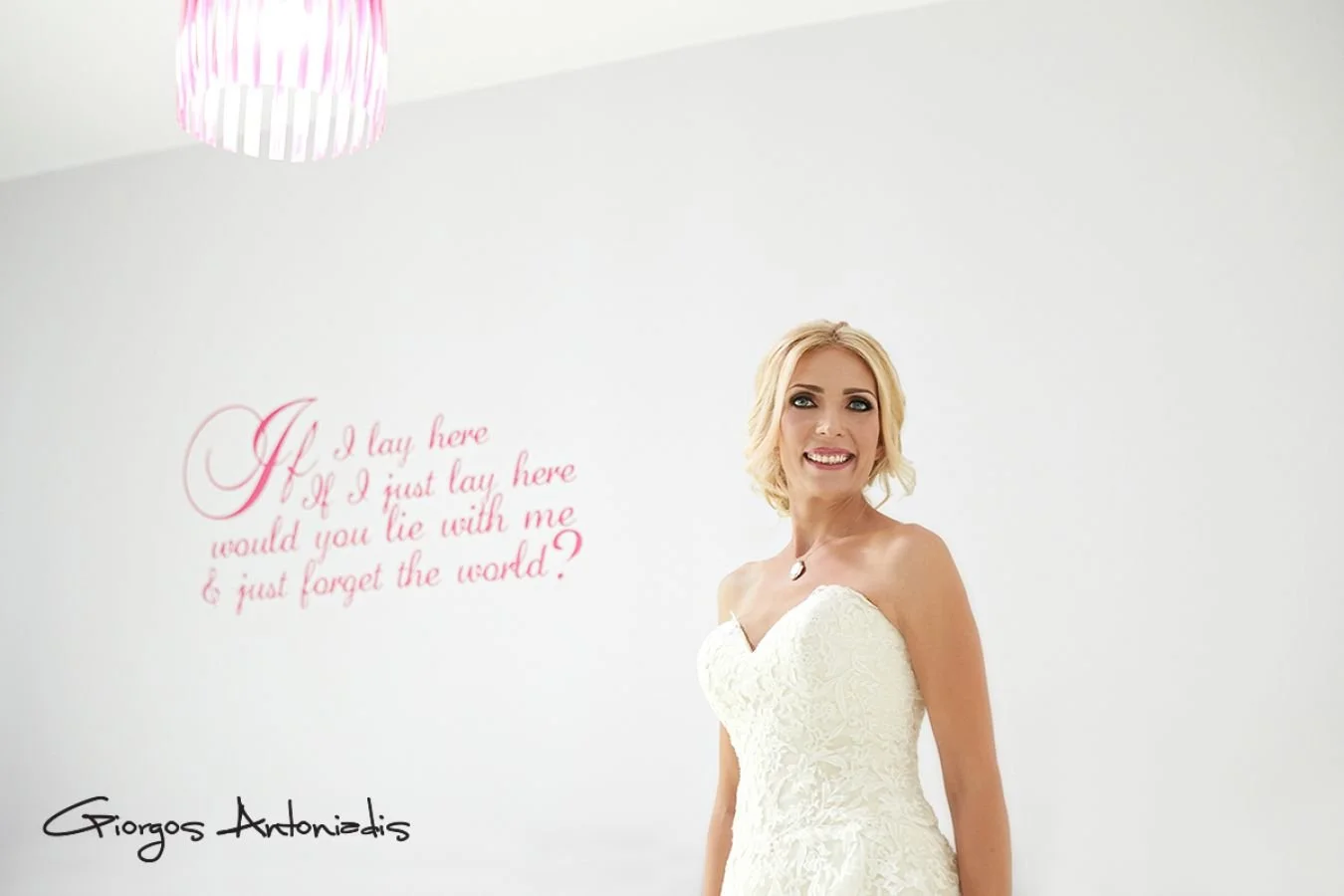 A smiling woman in a strapless white dress standing in front of a white wall with pink and black text. The pink text reads a romantic quote, and the black text appears to be a signature.
