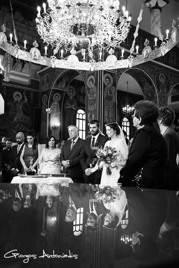 A black-and-white photo of a wedding ceremony inside a church, with a chandelier hanging from the ceiling. The bride and groom are standing together in front of a table, holding hands and facing an officiant. The bride is holding a bouquet, and all a