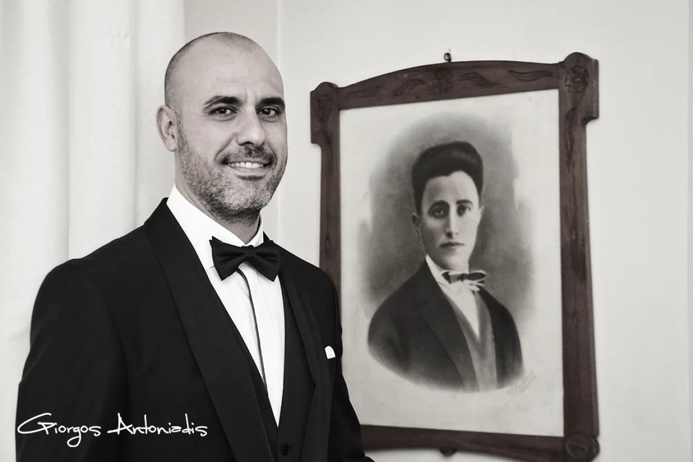 A man in a tuxedo with a bowtie standing next to a framed black and white portrait of a woman in vintage attire.