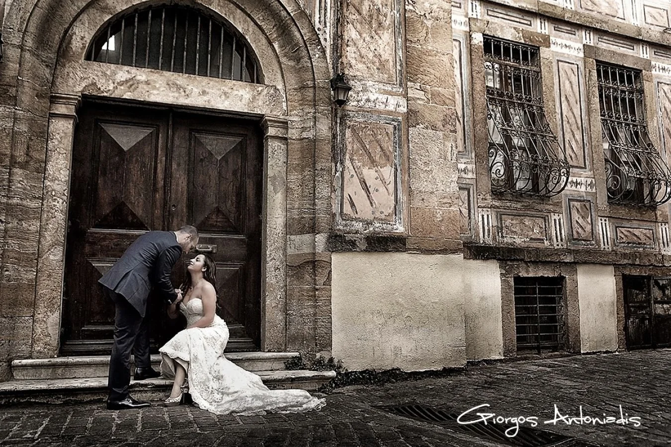 A man leaning over a woman in a white lace dress, sitting on steps outside a historic building with ornate stonework and iron window grilles. The scene appears romantic, with the man and woman engaging in an intimate moment.
