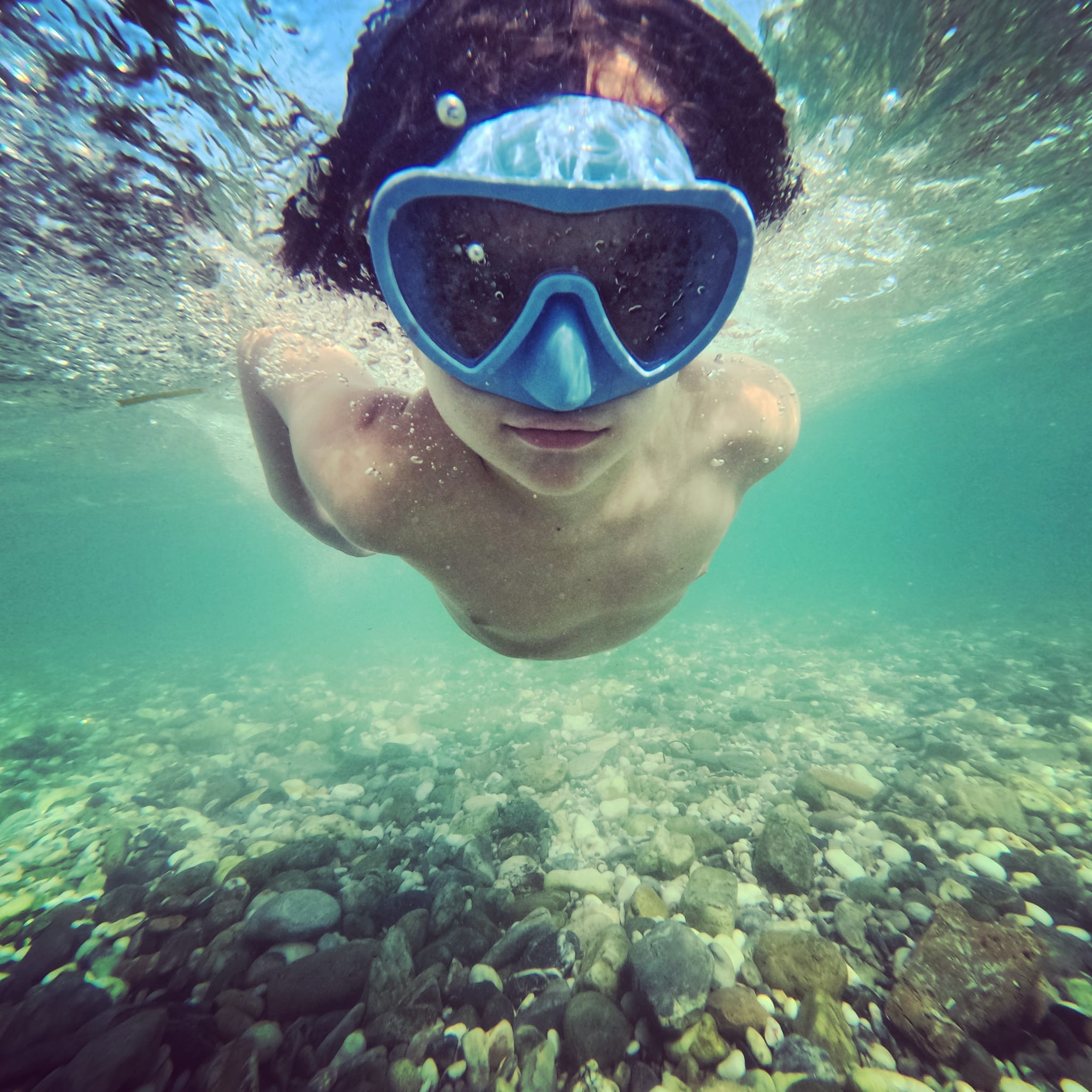 A person snorkeling underwater with a blue mask, swimming above a rocky riverbed.
