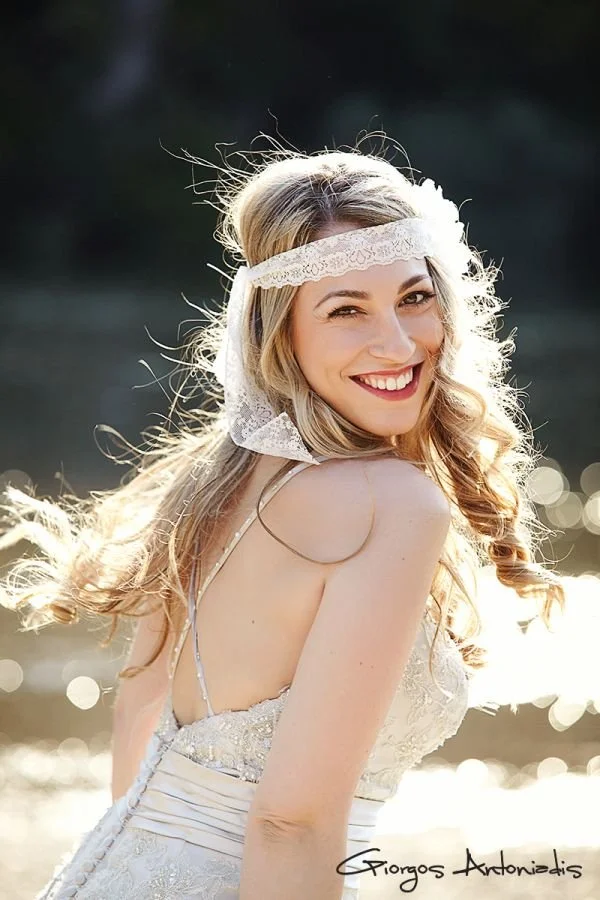 A smiling woman with long blonde hair wearing a lace headband and a white dress outdoors with sunlight and water in the background.