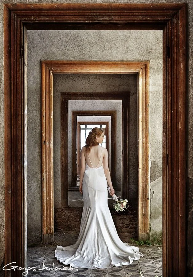 A woman in a long white wedding dress holding a bouquet, standing in an abandoned building with multiple doorways creating a tunnel effect.