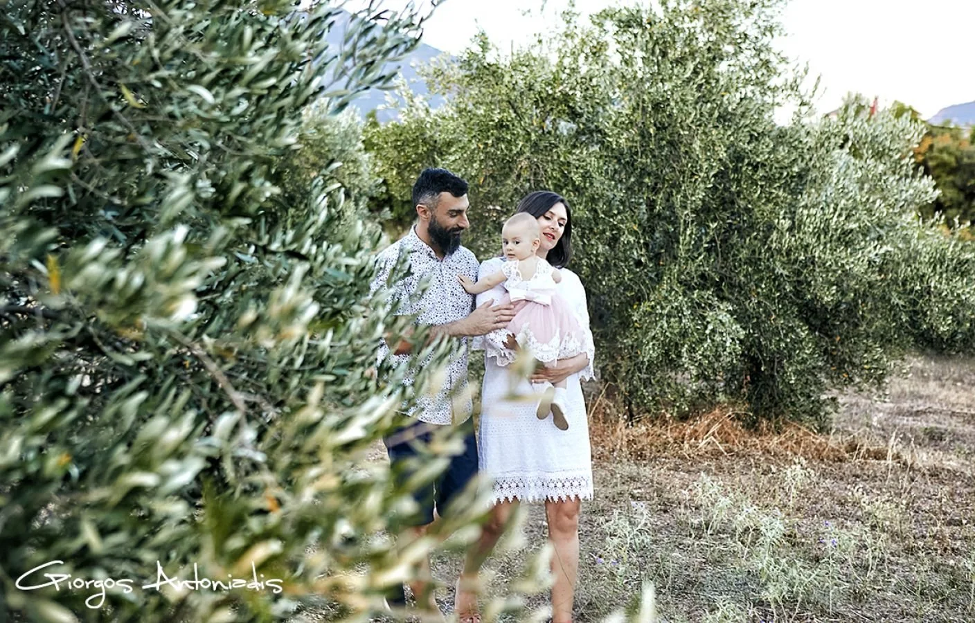 A family walking through a natural landscape with green bushes and trees, with a man holding a young girl and a woman walking beside them.