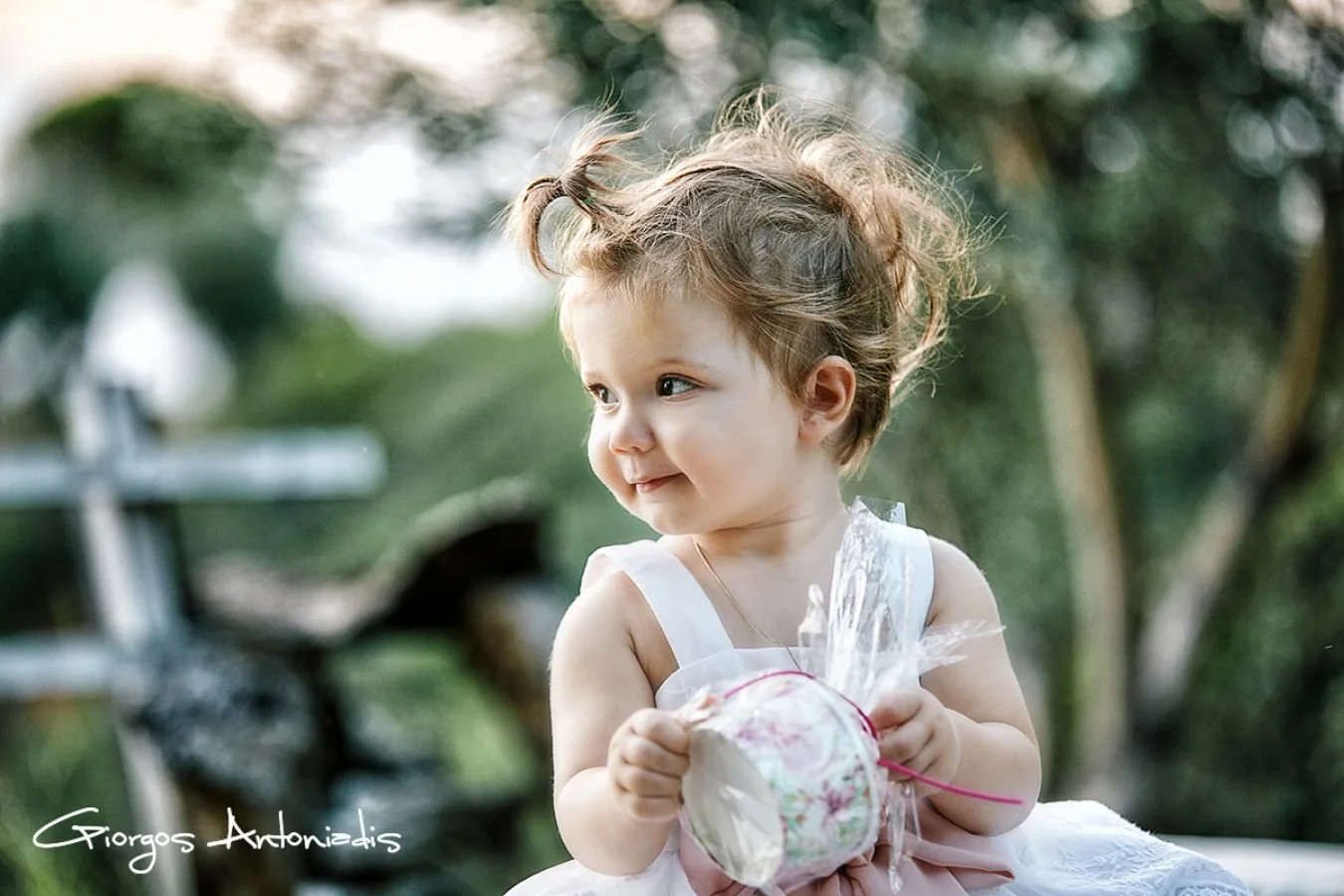 A young girl with curly hair in a ponytail, wearing a white dress with pink accents, sitting outdoors with a soft smile, holding a decorated Easter egg wrapped in clear plastic with feathers inside, with blurred greenery and an outdoor fountain or sc