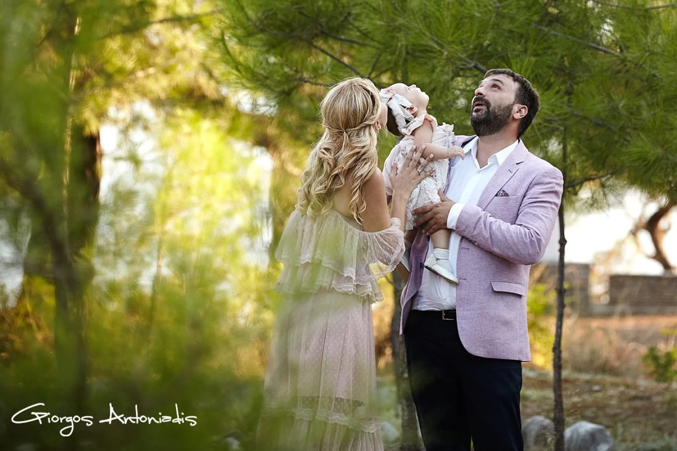 A family of three outdoors, with a woman, a man, and a small girl, surrounded by green trees, enjoying a happy moment together.