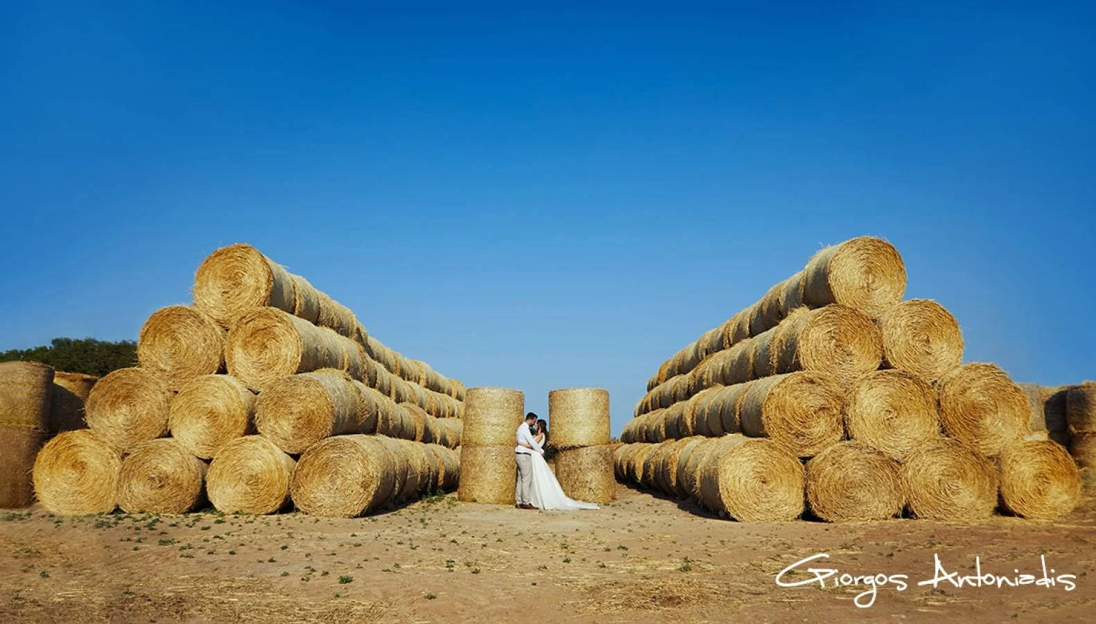 A couple dressed in white wedding attire embracing each other in a field with large stacks of hay bales arranged in triangular formations on either side. Clear blue sky in background.
