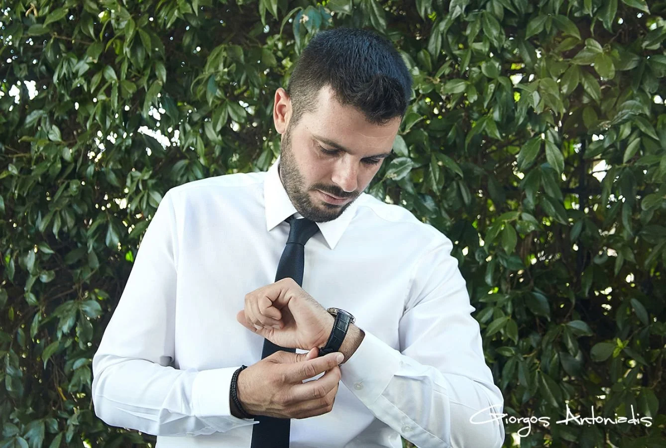 A man in a white shirt and black tie checks the time on his watch outdoors against a background of green leaves.