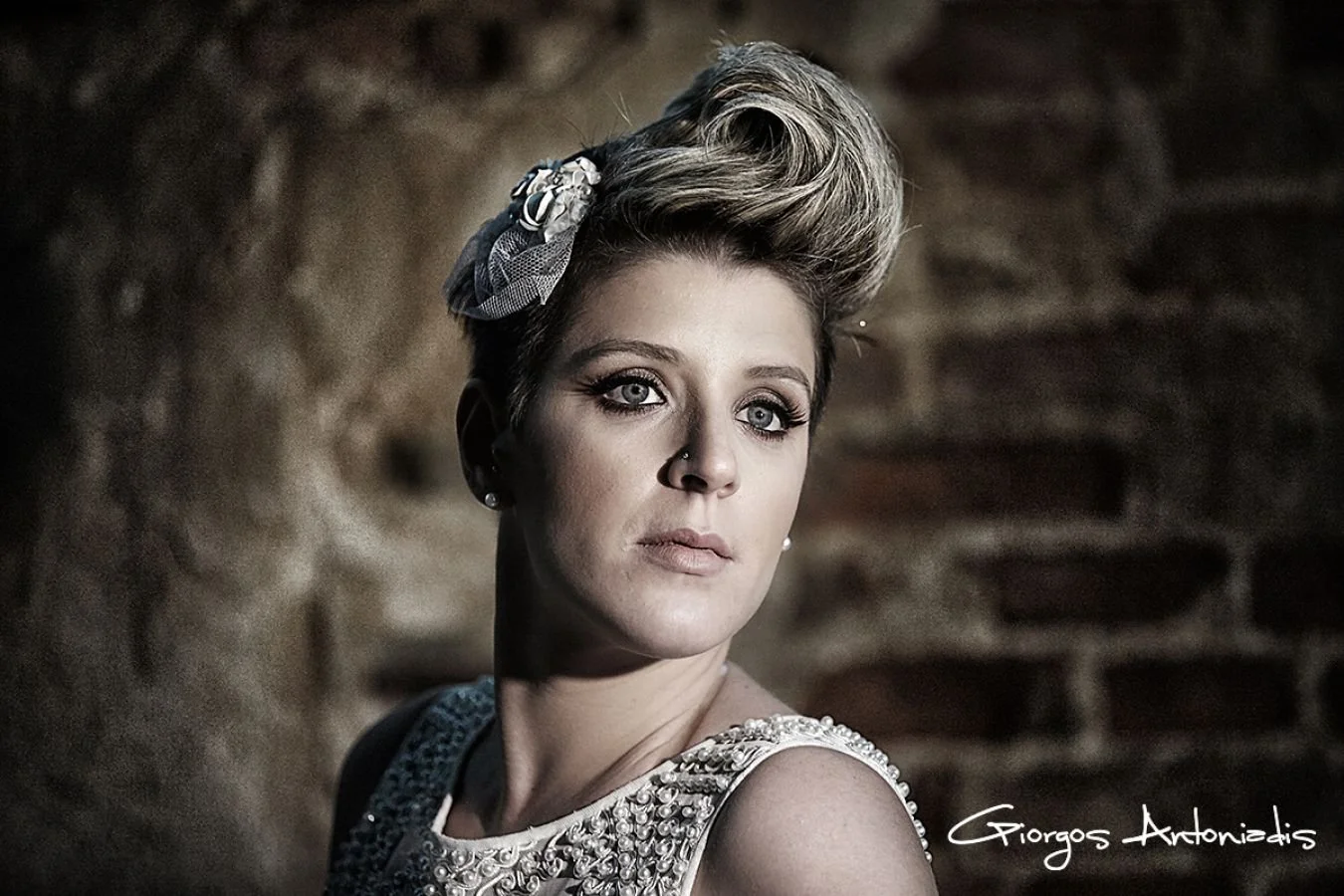 A woman with short, styled blonde hair and blue eyes, wearing pearl earrings and a sleeveless dress with decorative beads, stands against a blurred brick wall background.