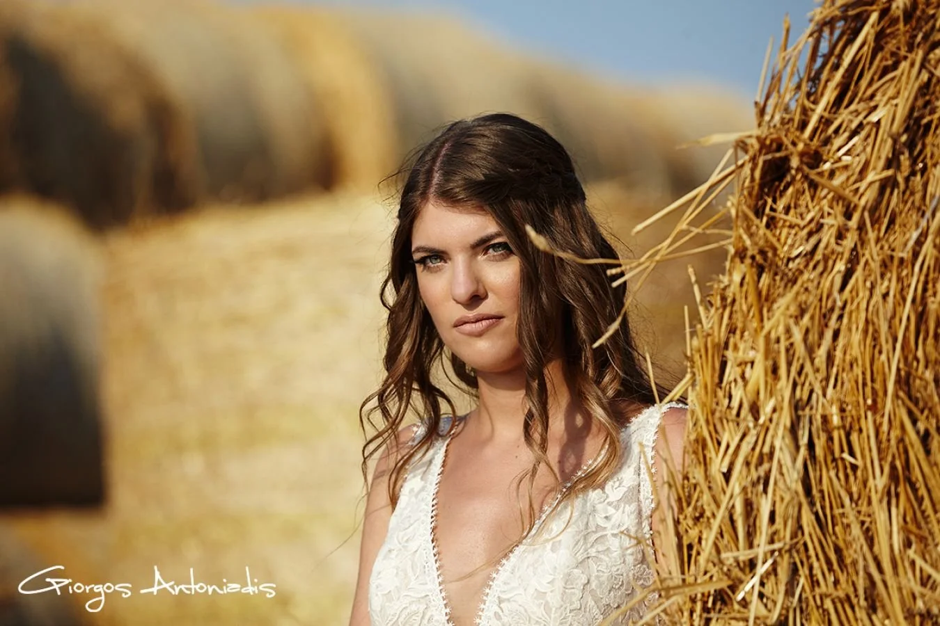 A woman with wavy brown hair and light skin standing next to a haystack in a field during daytime, wearing a white lace top.