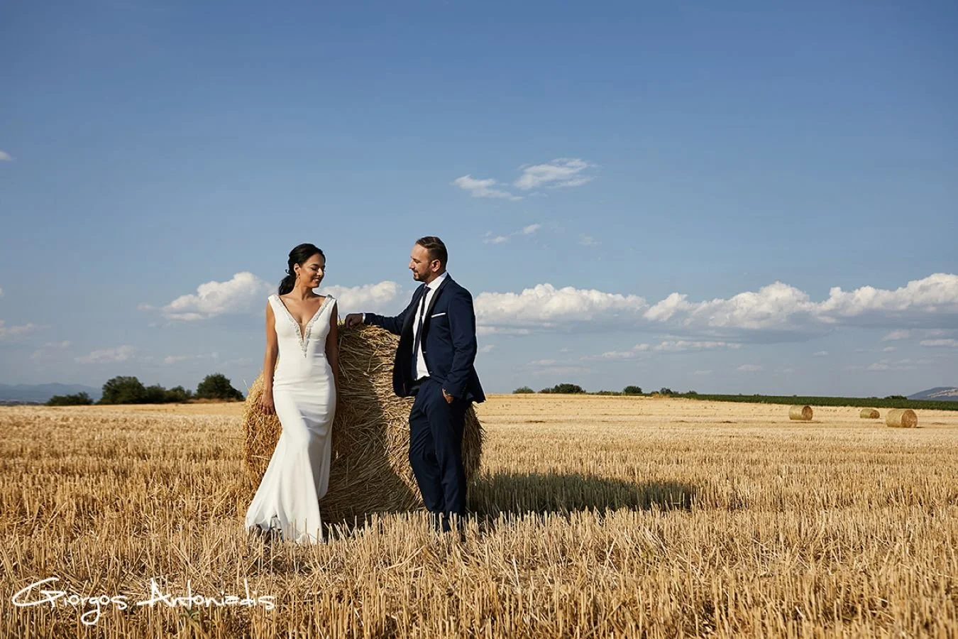 A man in a navy suit and a woman in a white wedding dress standing next to a large hay bale in a wheat field under a blue sky with scattered clouds.