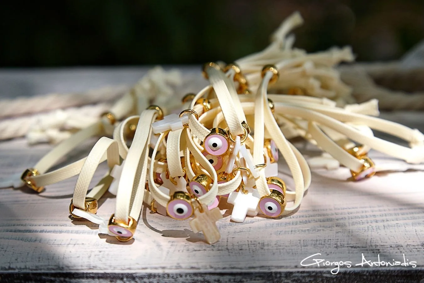 A pile of cream-colored wristbands with pink and white eye charms and small wooden crosses, adorned with gold accents, scattered on a surface.