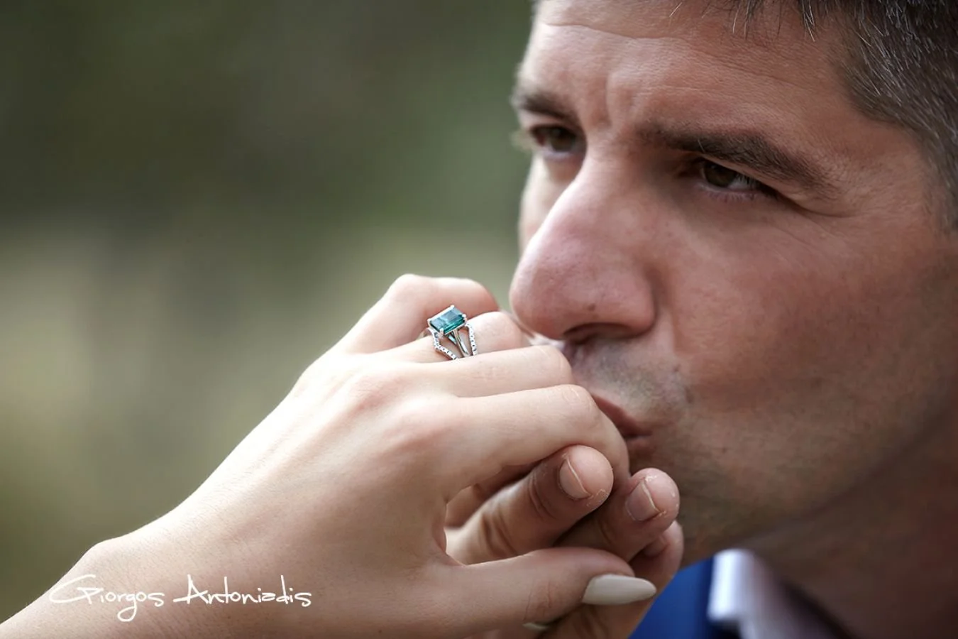 A man is deep in thought with a woman kissing his hand, wearing a ring, and his eyes gazing away, outdoors with a blurred background.