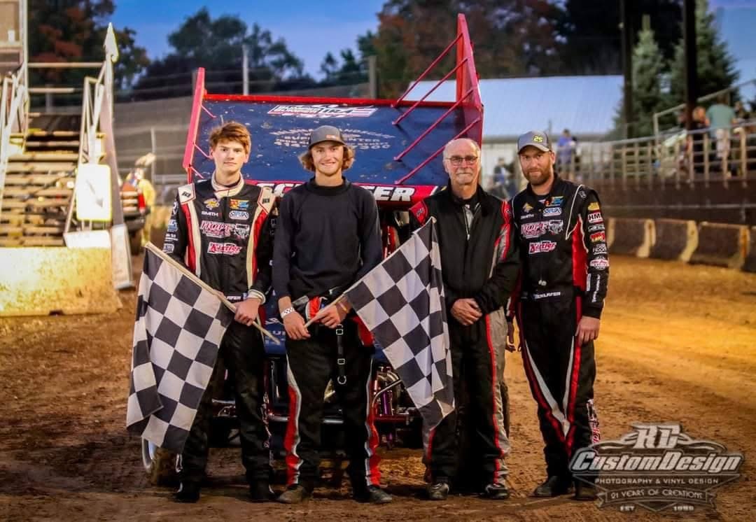 Group of four people including two race car drivers in racing suits, one young man in casual clothes, and an older man, standing in a dirt race track with a sprint car behind them. The two race car drivers are holding checkered flags.
