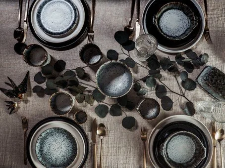 Bunte Keramik-Tassen und -Schalen auf einem Holztablett in einem Geschäft, im Hintergrund sind ungeordnete Regale mit weiteren Produkten.