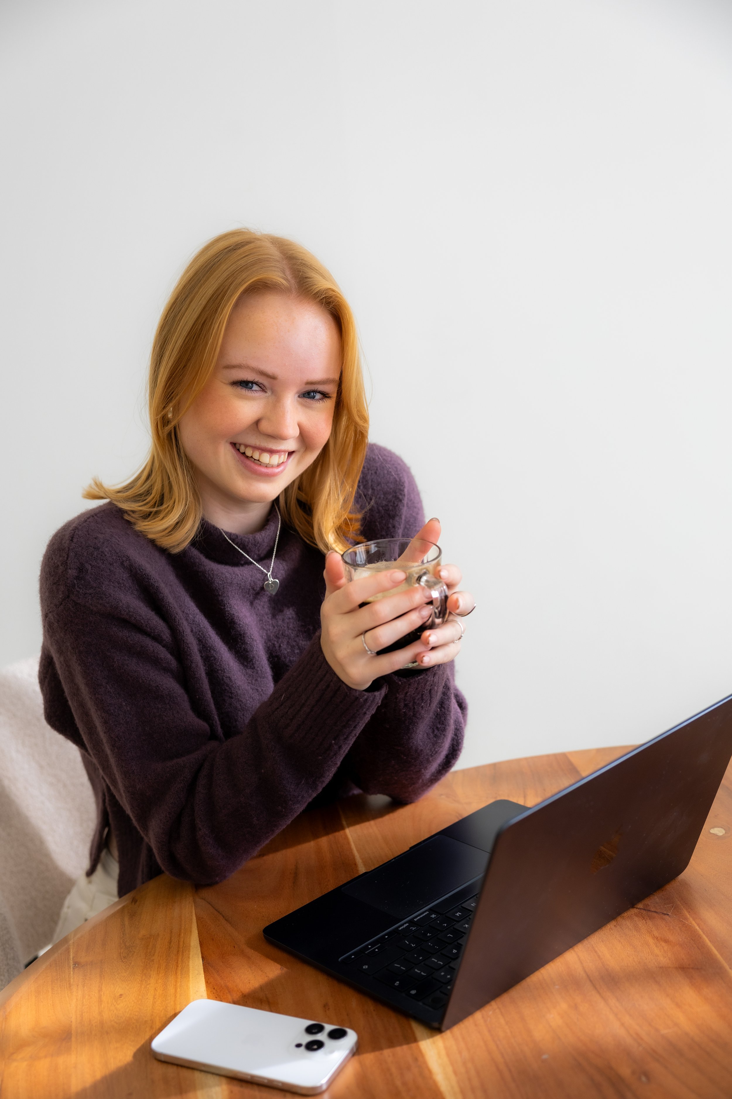 Lachende vrouw met rood haar houdt een kopje koffie of thee vast, zittend voor een laptop op een houten tafel, met een witte smartphone naast haar.