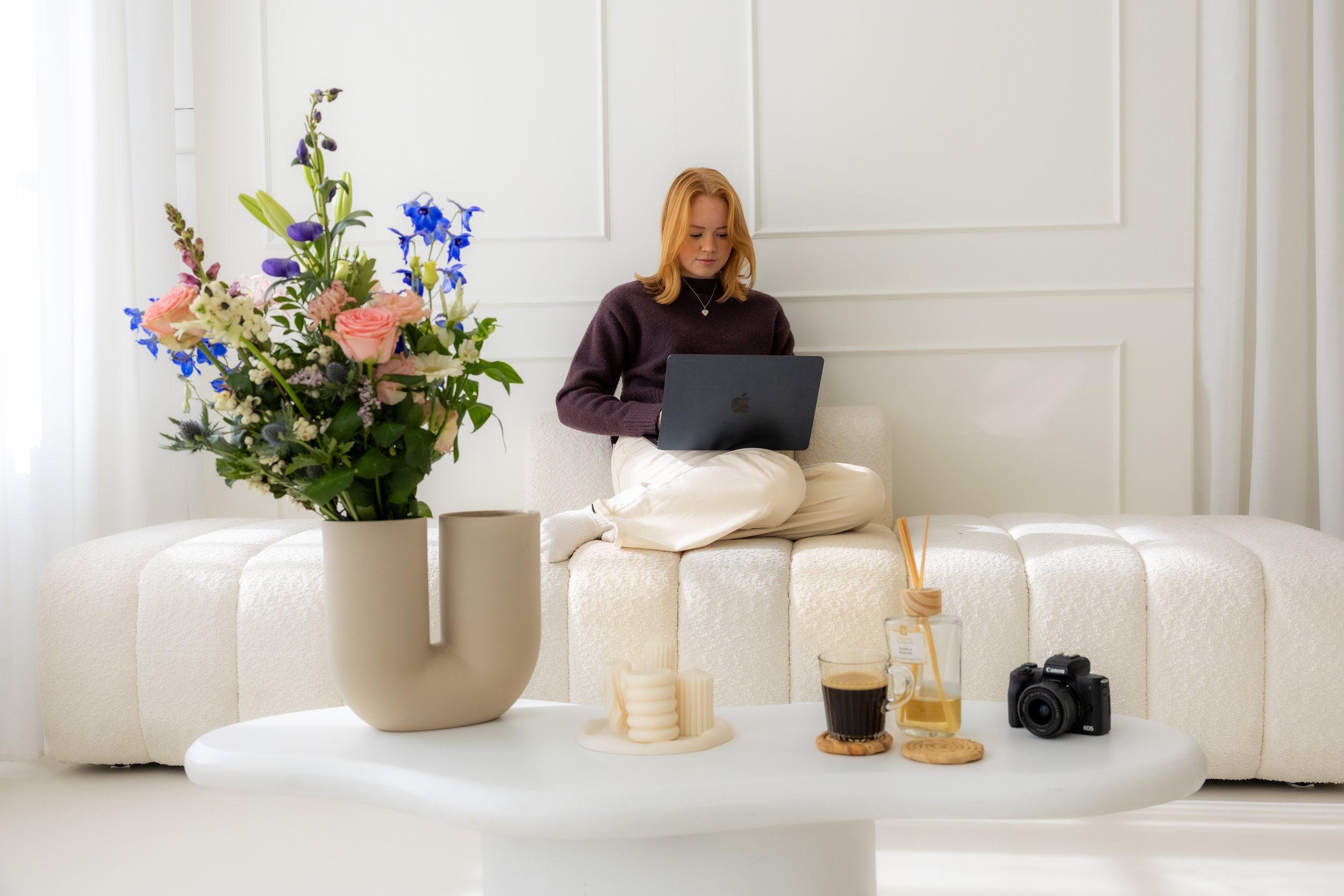 Vrouw zit op een bank met een laptop op schoot, omringd door bloemen, kaarsen, koffie en een camera in een lichte, rustige ruimte.