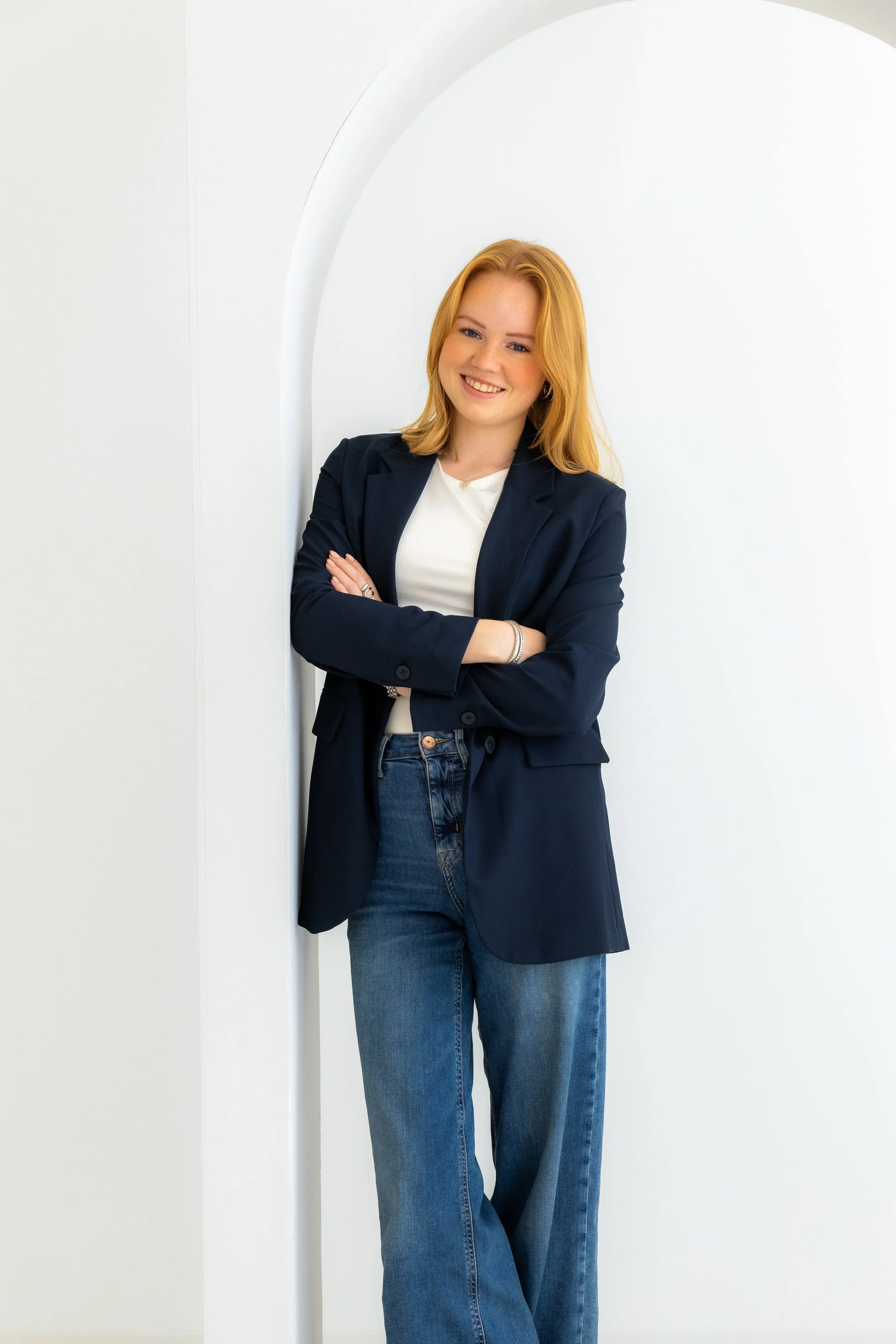 Warm geklede vrouw met rode haar die lacht en met gekruiste armen tegen een witte muur leunt.