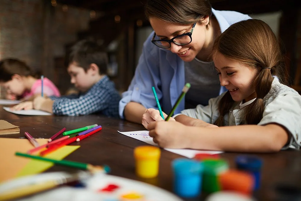 A woman and girl drawing and coloring at a table with colored markers and paper, with children in the background.