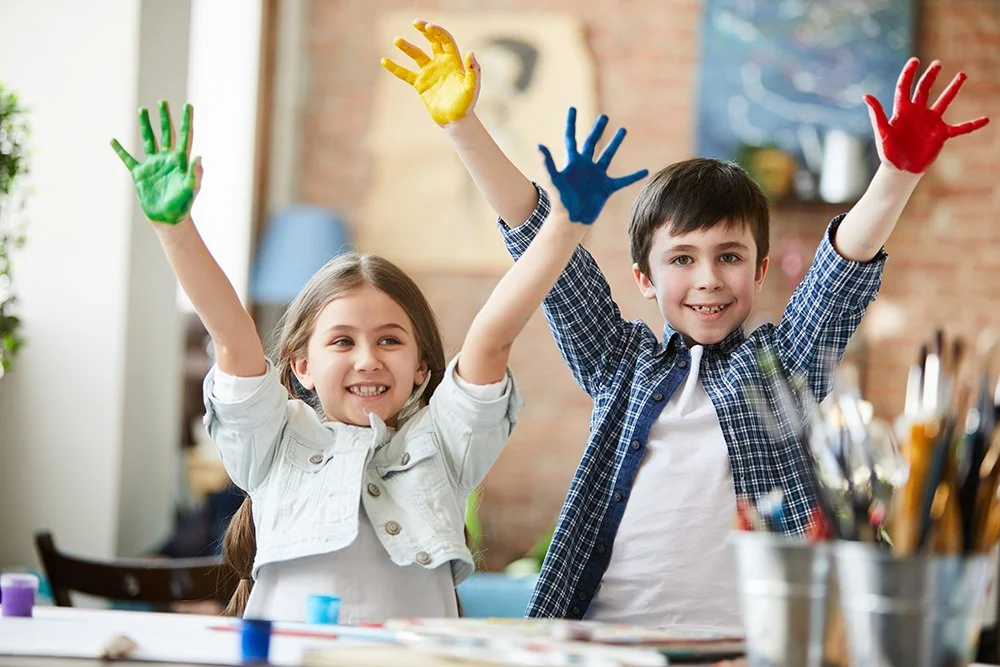 Two kids, a girl and a boy, with painted hands, raising their hands in a cheerful art classroom.