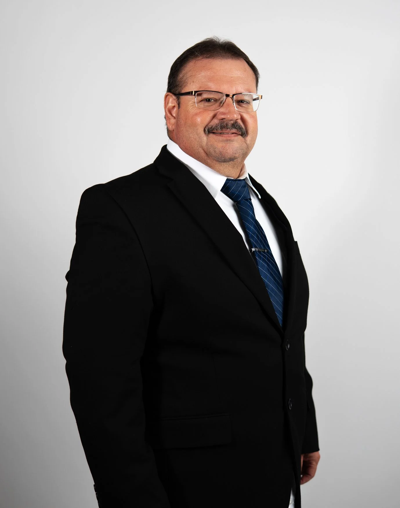A middle-aged man with glasses, a mustache, wearing a black suit, white shirt, and navy blue tie, standing against a plain light gray background.