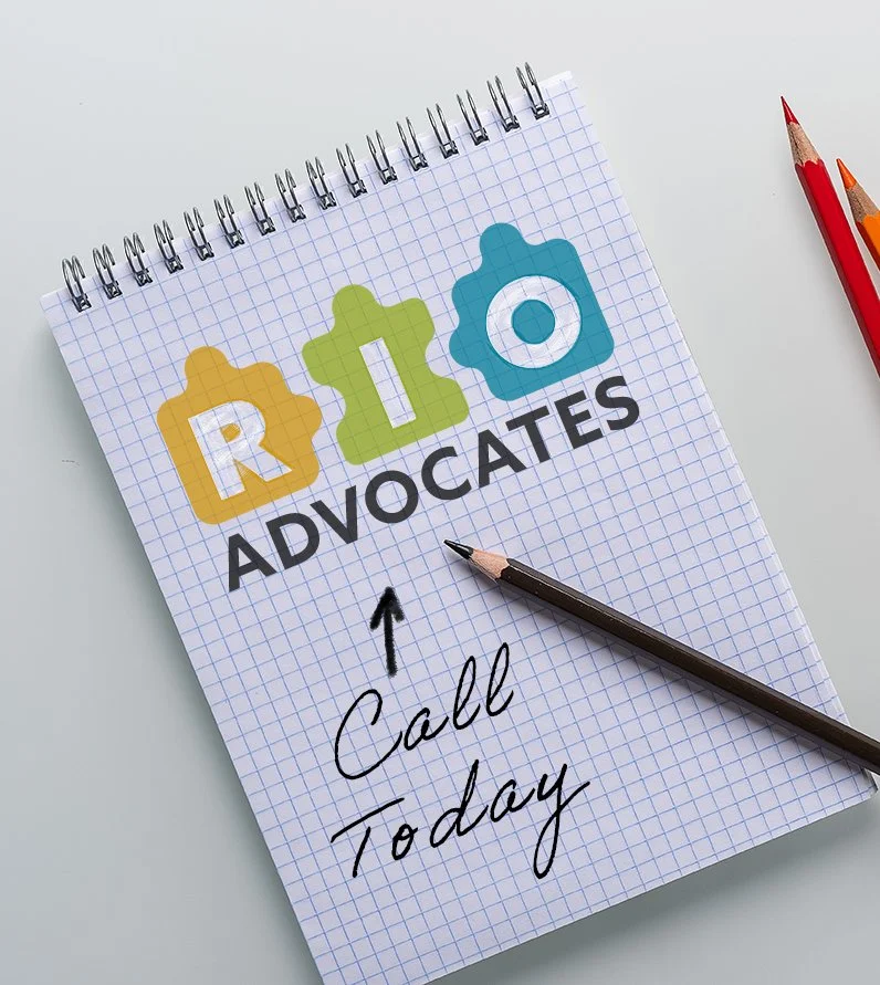 A notepad with the logo 'RIO Advocates' on it, with colorful puzzle pieces forming the letters R, I, and O. Below the logo, there is handwritten text with an arrow pointing up that says 'Call Today'. There are pens nearby on a white surface.