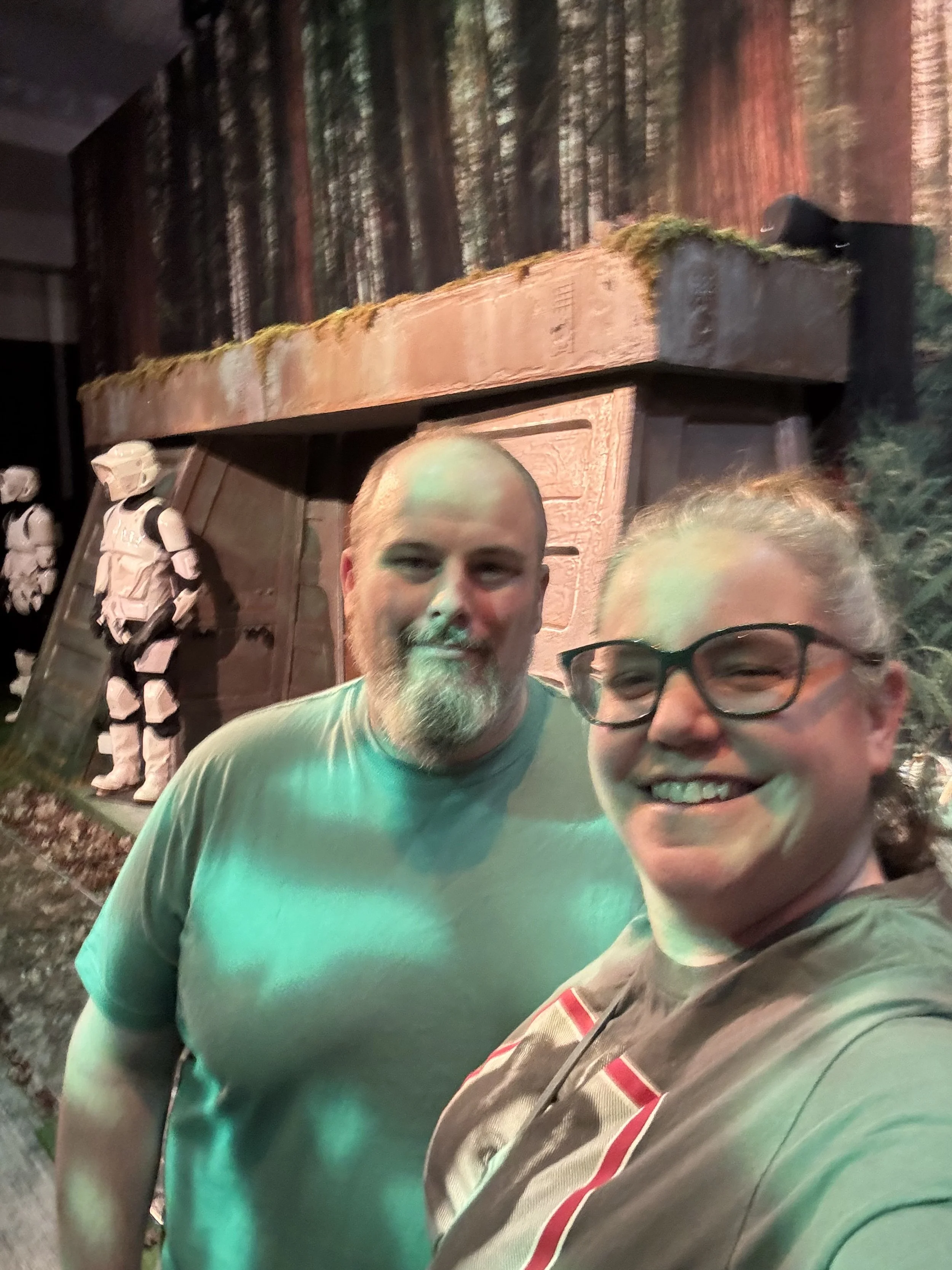 Two people taking a selfie in front of a Star Wars display, which includes a model of a stormtrooper and a landscape background resembling a forest.