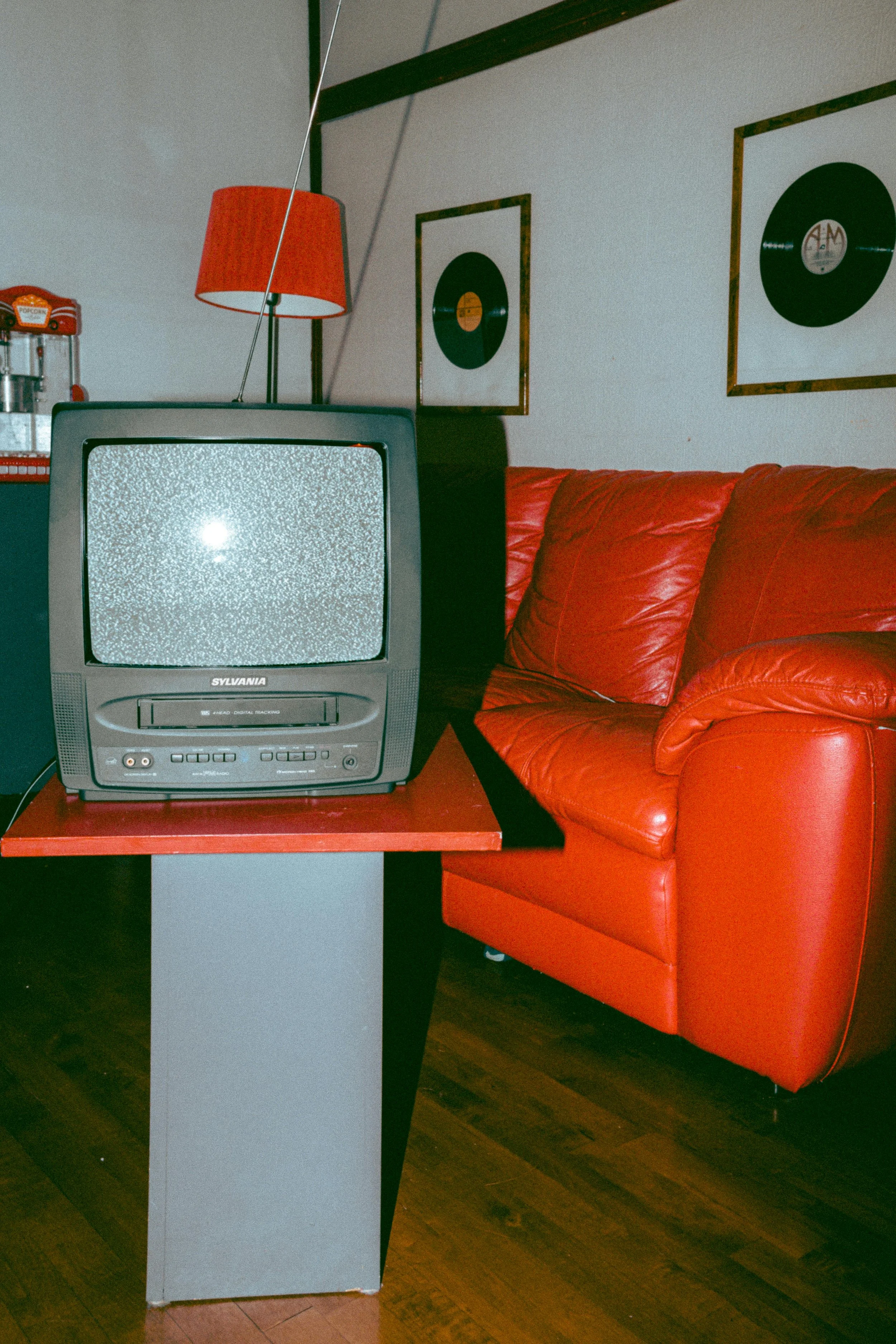 Vintage television set on a table next to a red couch in a living room with framed records on the wall and a floor lamp.