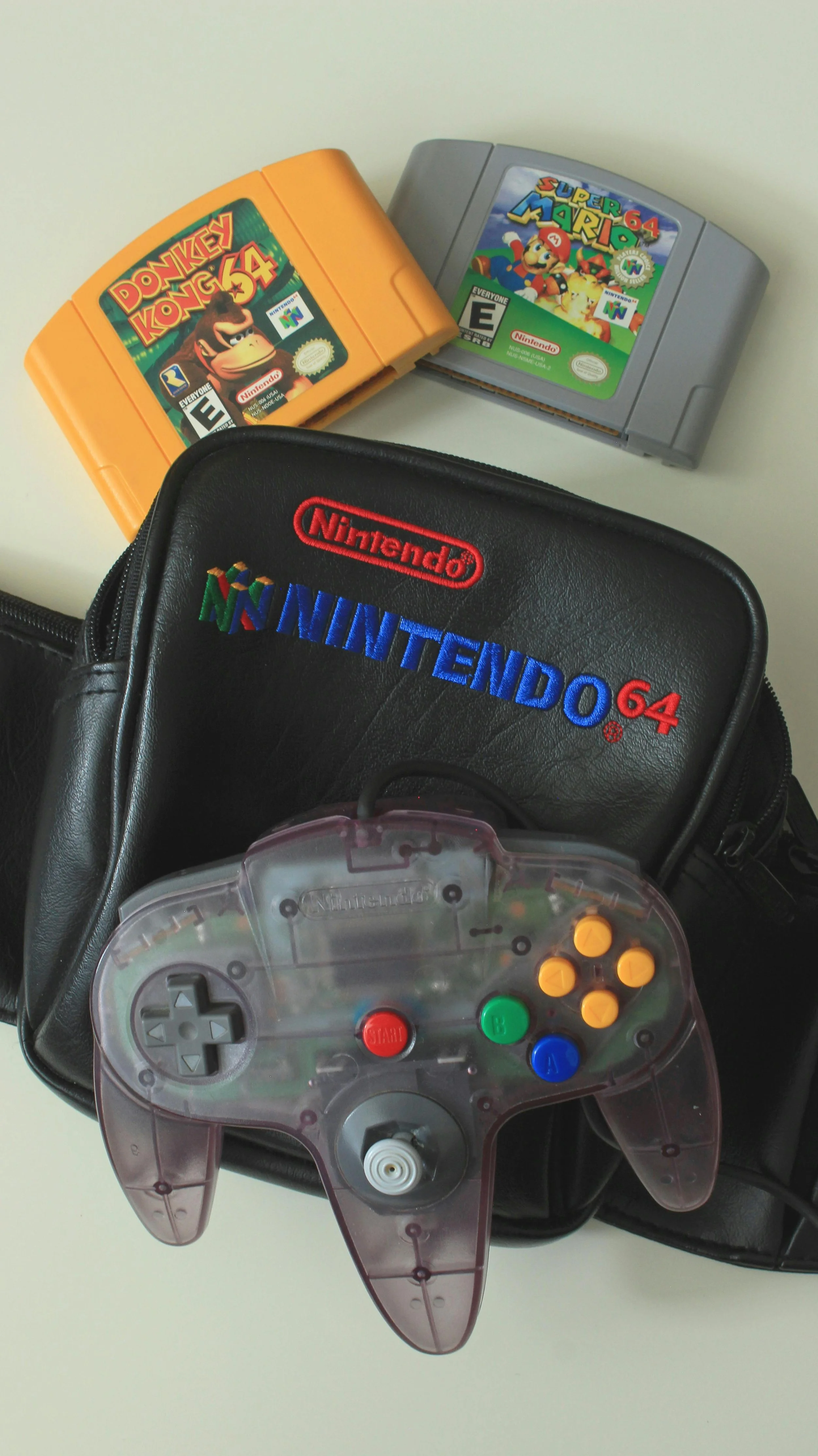 A Nintendo 64 black carrying case with the Nintendo logo and Nintendo 64 logo embroidered on it, two Nintendo 64 game cartridges (Donkey Kong 64 in the yellow case and Super Mario 64 in the gray case) placed on a light-colored surface, and a transparent Nintendo 64 controller with multi-colored buttons.