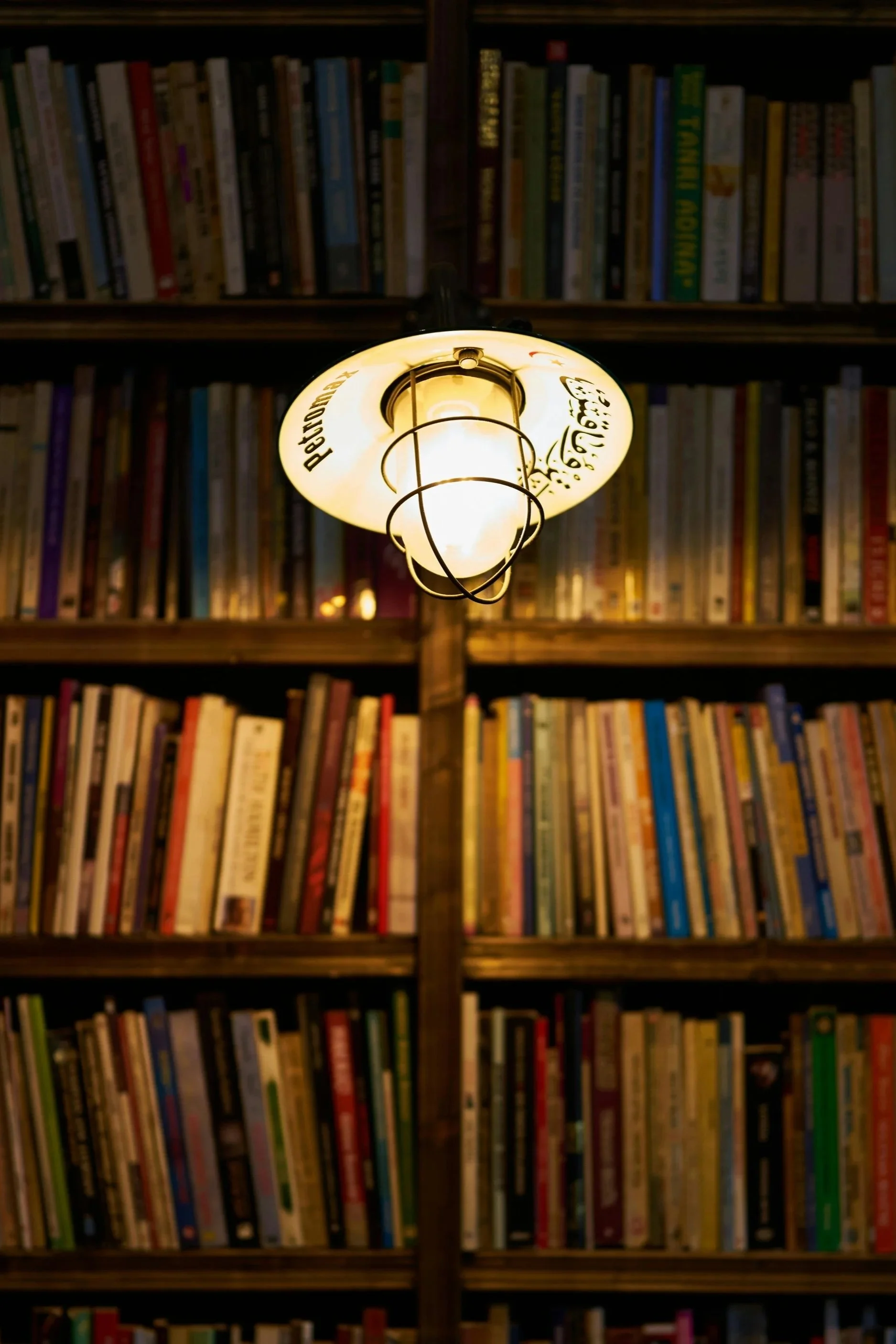 A ceiling light fixture with a yellowish glow, decorated with black script and symbols, hangs in front of a background of bookshelves filled with colorful books.